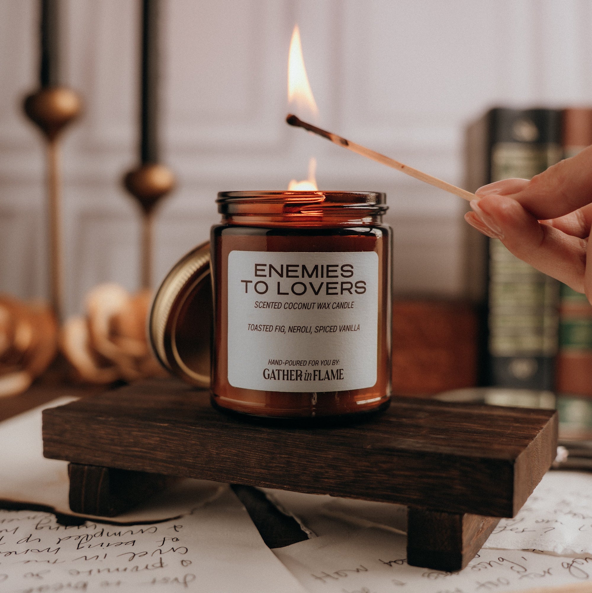 Candle labeled 'Enemies to Lovers' being lit on a wooden stand with books and candles in the background.