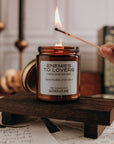 Candle labeled 'Enemies to Lovers' being lit on a wooden stand with books and candles in the background.