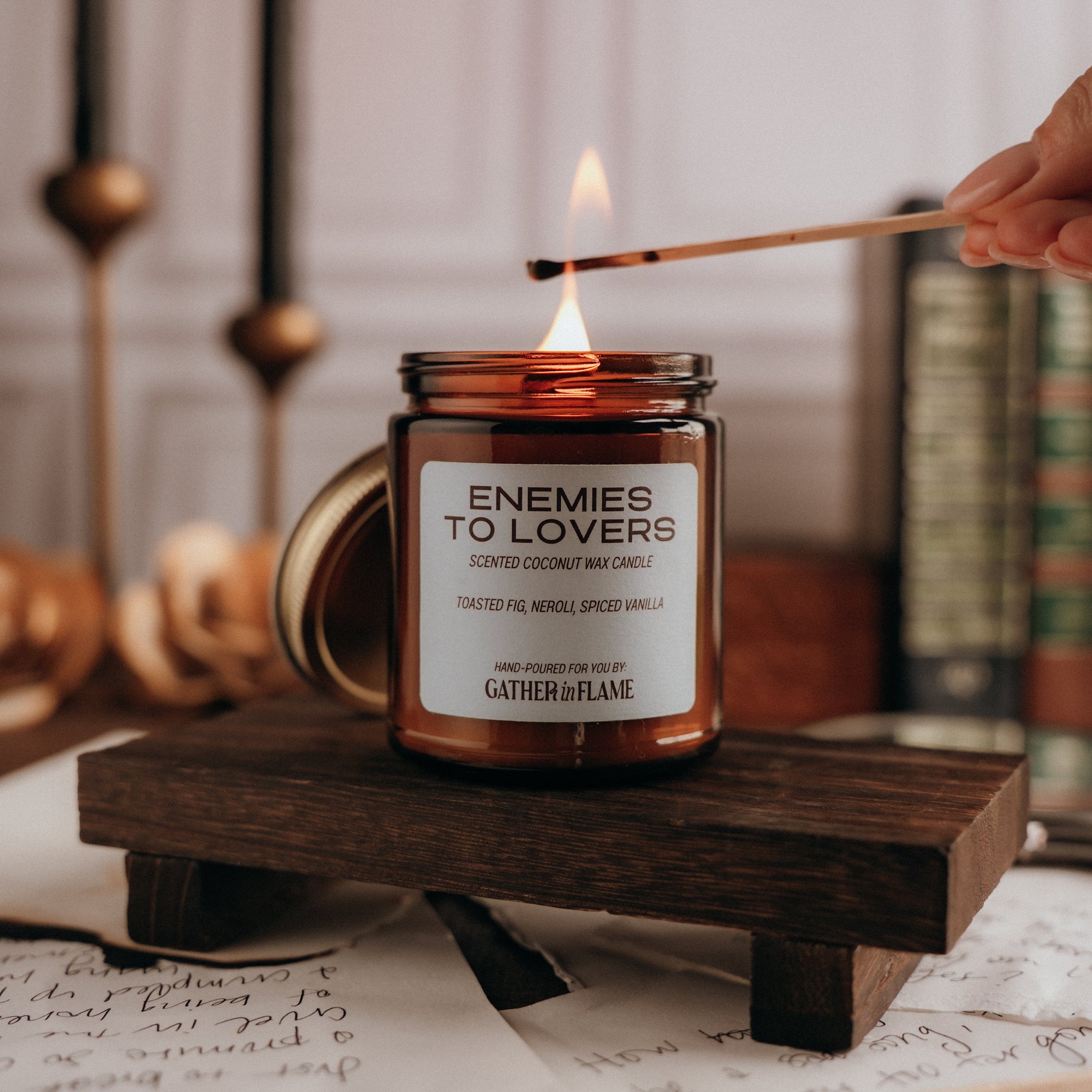Candle labeled 'Enemies to Lovers' being lit on a wooden stand with books and candles in the background.