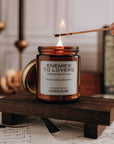 Candle labeled 'Enemies to Lovers' being lit on a wooden stand with books and candles in the background.