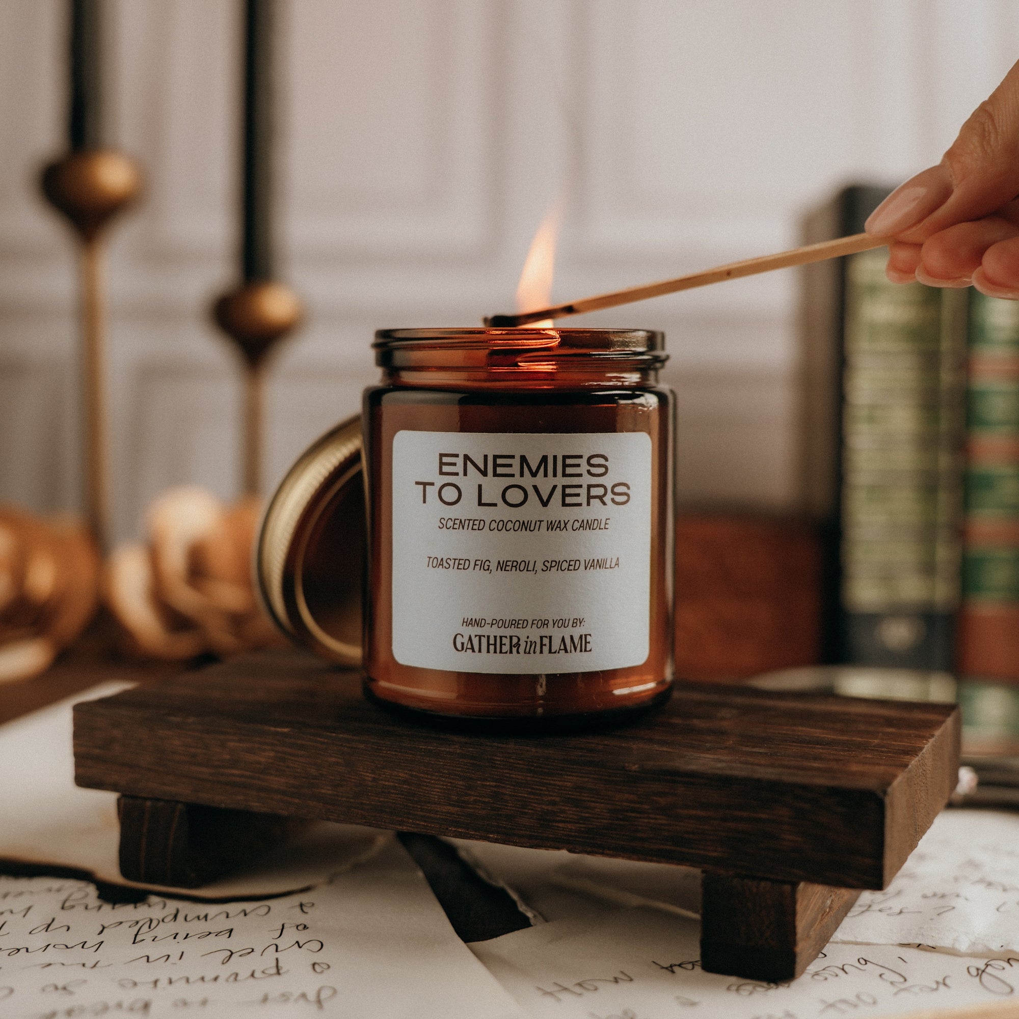 Candle labeled 'Enemies to Lovers' being lit on a wooden stand with books and candles in the background.