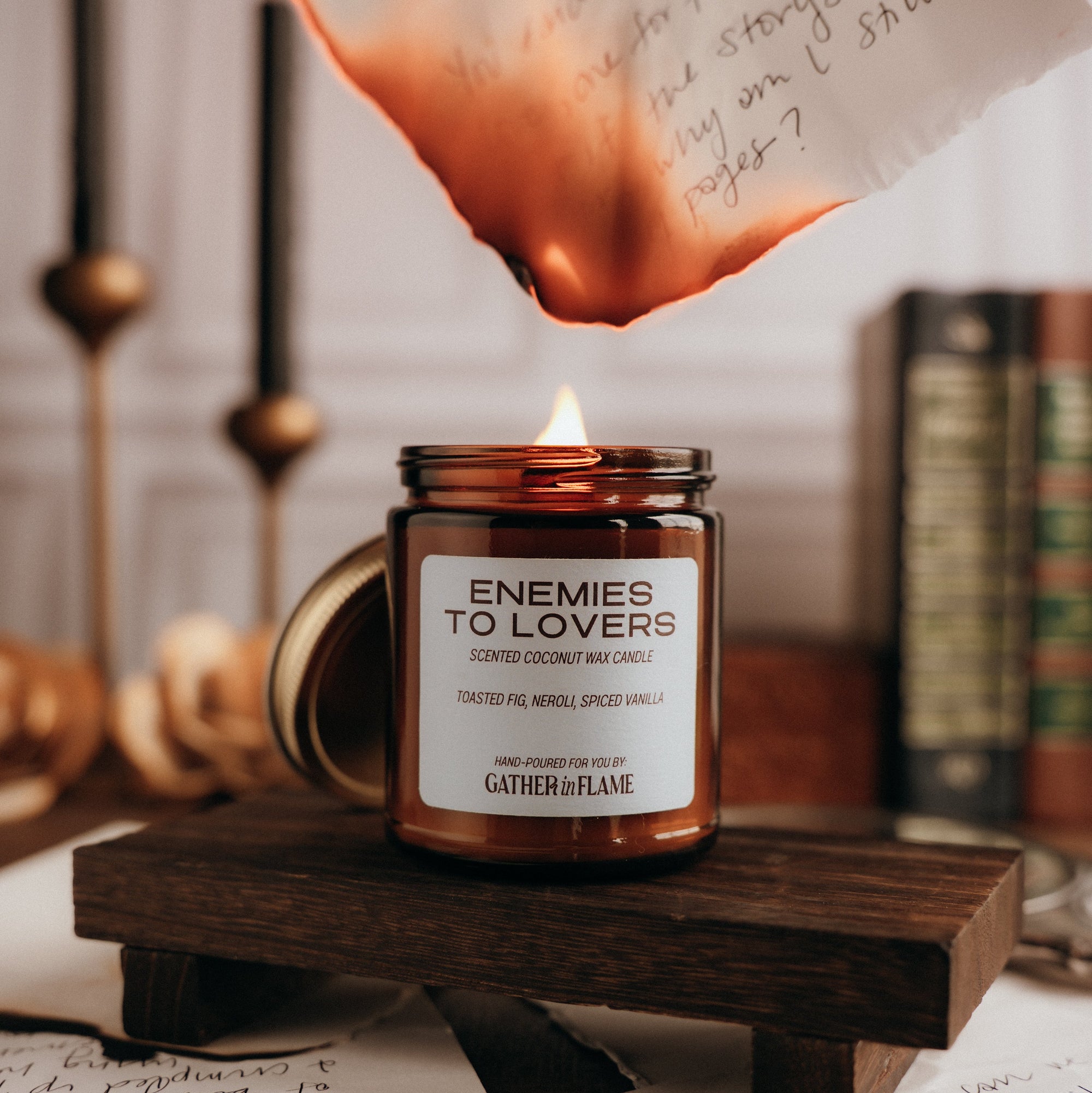 Candle labeled 'Enemies to Lovers' on a wooden stand with books and papers in the background and note burning above it.