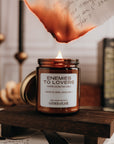 Candle labeled 'Enemies to Lovers' on a wooden stand with books and papers in the background and note burning above it.
