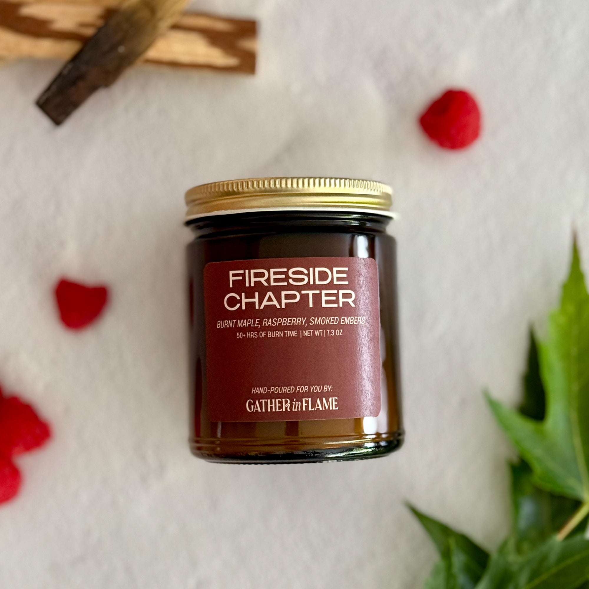 Candle jar labeled 'Fireside Chapter' by Gather the Flame on a textured white background with raspberries, wood pieces and green leaves.