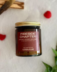 Candle jar labeled 'Fireside Chapter' by Gather the Flame on a textured white background with raspberries, wood pieces and green leaves.