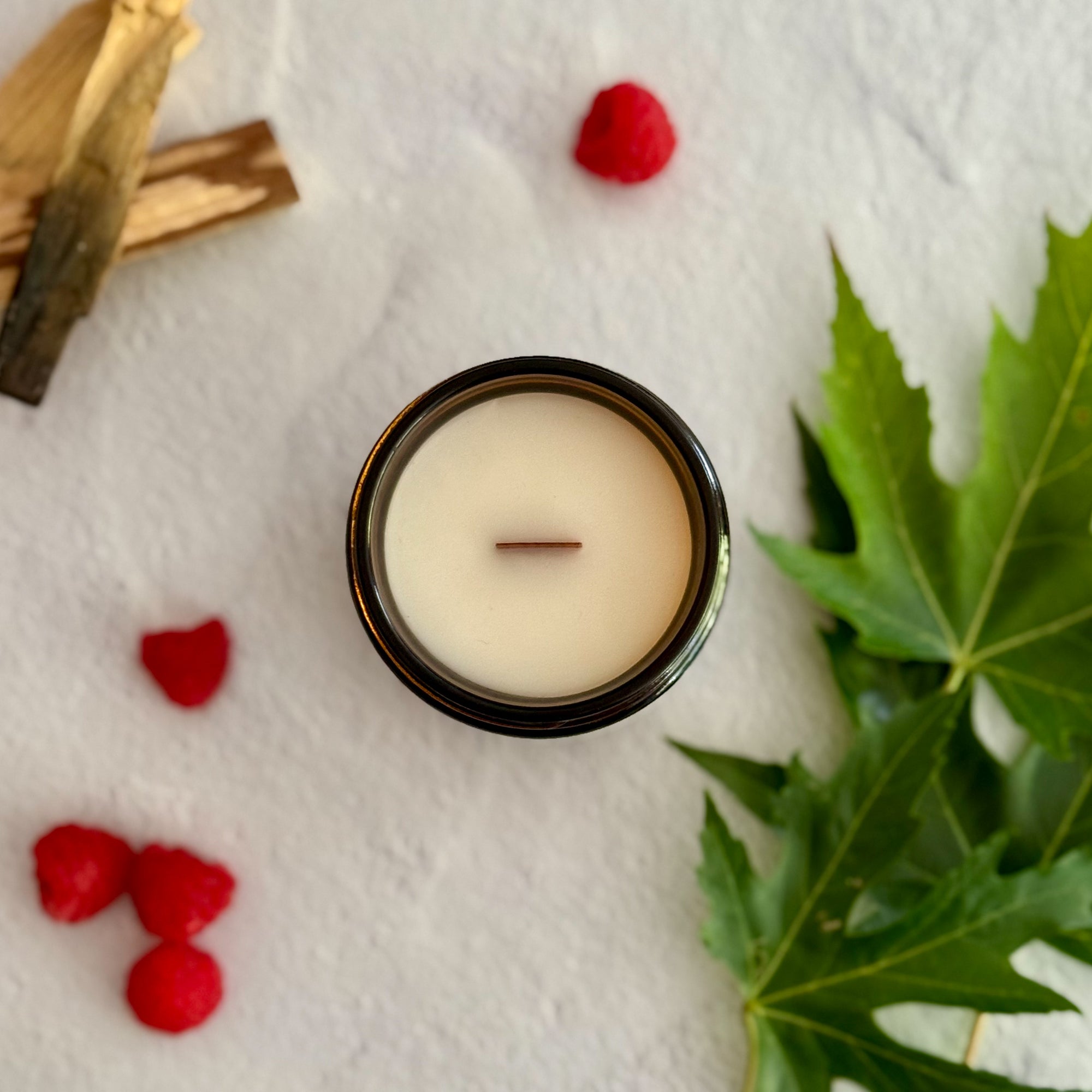 Topdown view of candle with wood wick surrounded by raspberries, wood pieces and green leaves on a white background