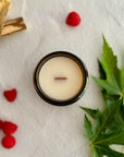 Topdown view of candle with wood wick surrounded by raspberries, wood pieces and green leaves on a white background