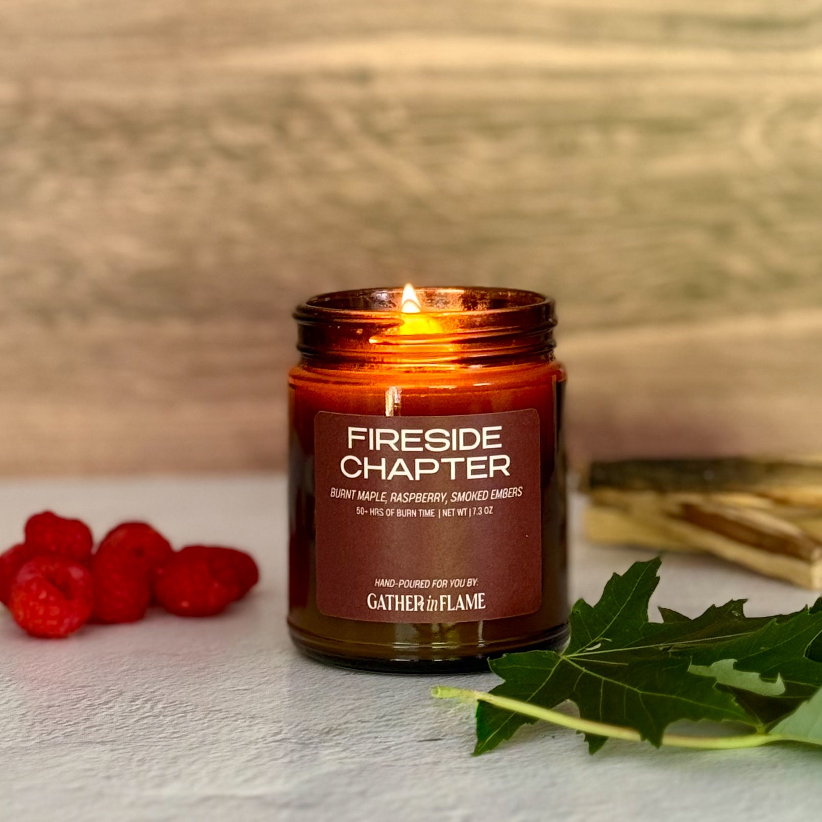 Lit candle labeled 'Fireside Chapter' with raspberries and leaves on a wooden surface