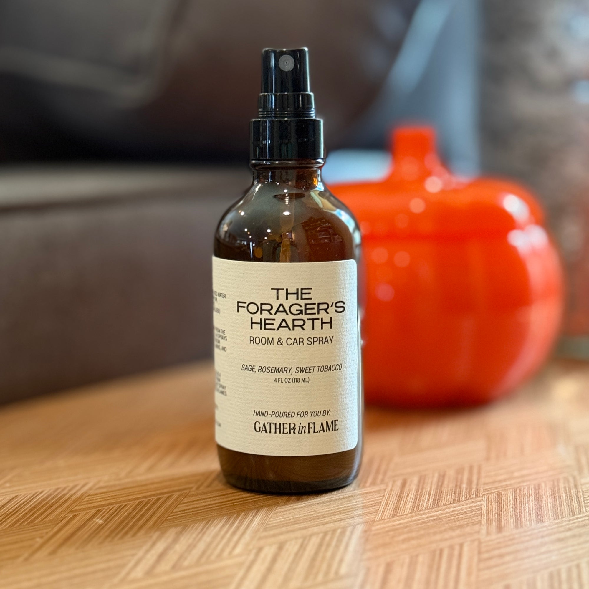 Bottle of 'The Forager's Hearth' room and car spray on a wooden surface with a blurred pumpkin in the background.