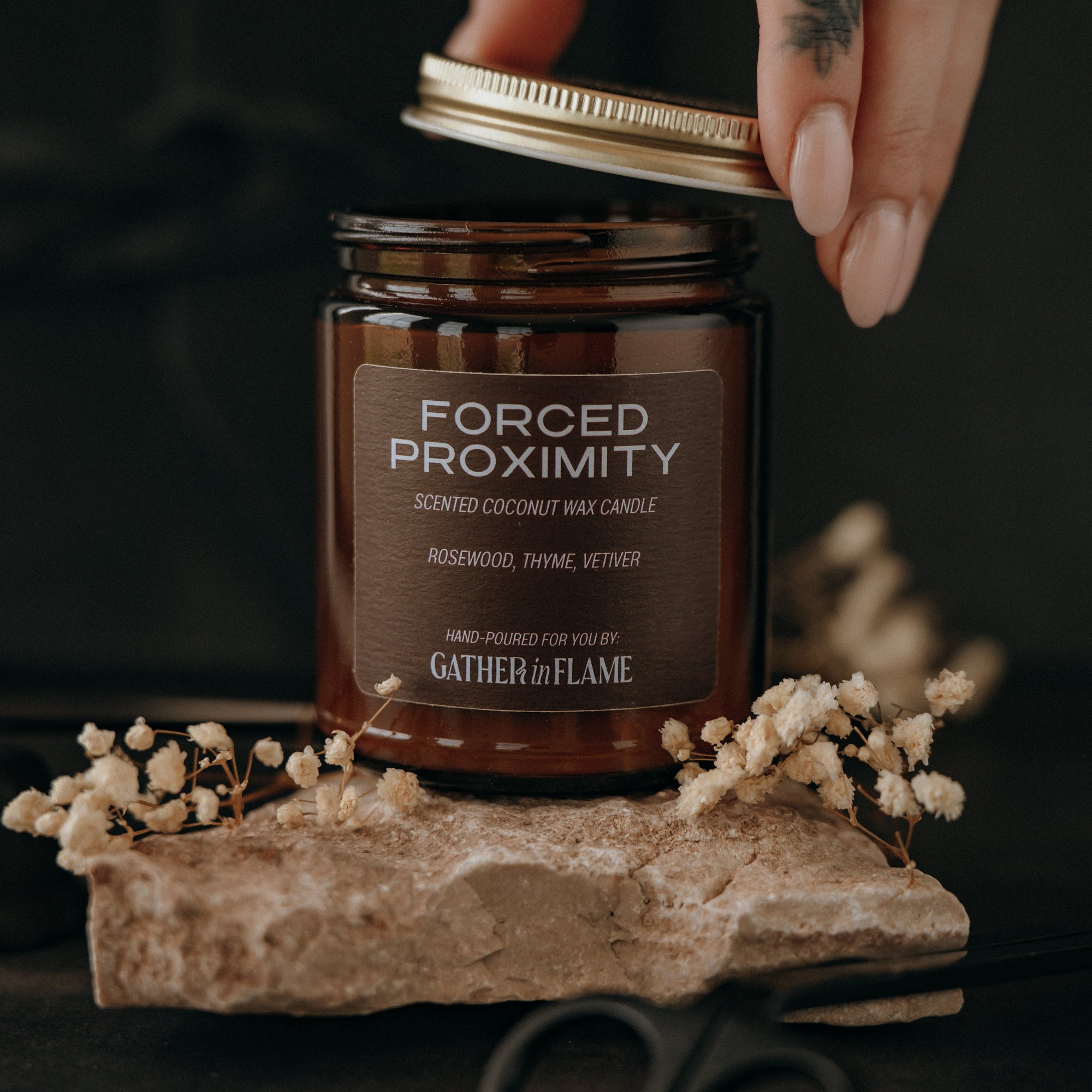 Jar of 'Forced Proximity' candle held by a hand with a tattoo, on a dark background.