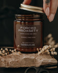 Jar of 'Forced Proximity' candle held by a hand with a tattoo, on a dark background.