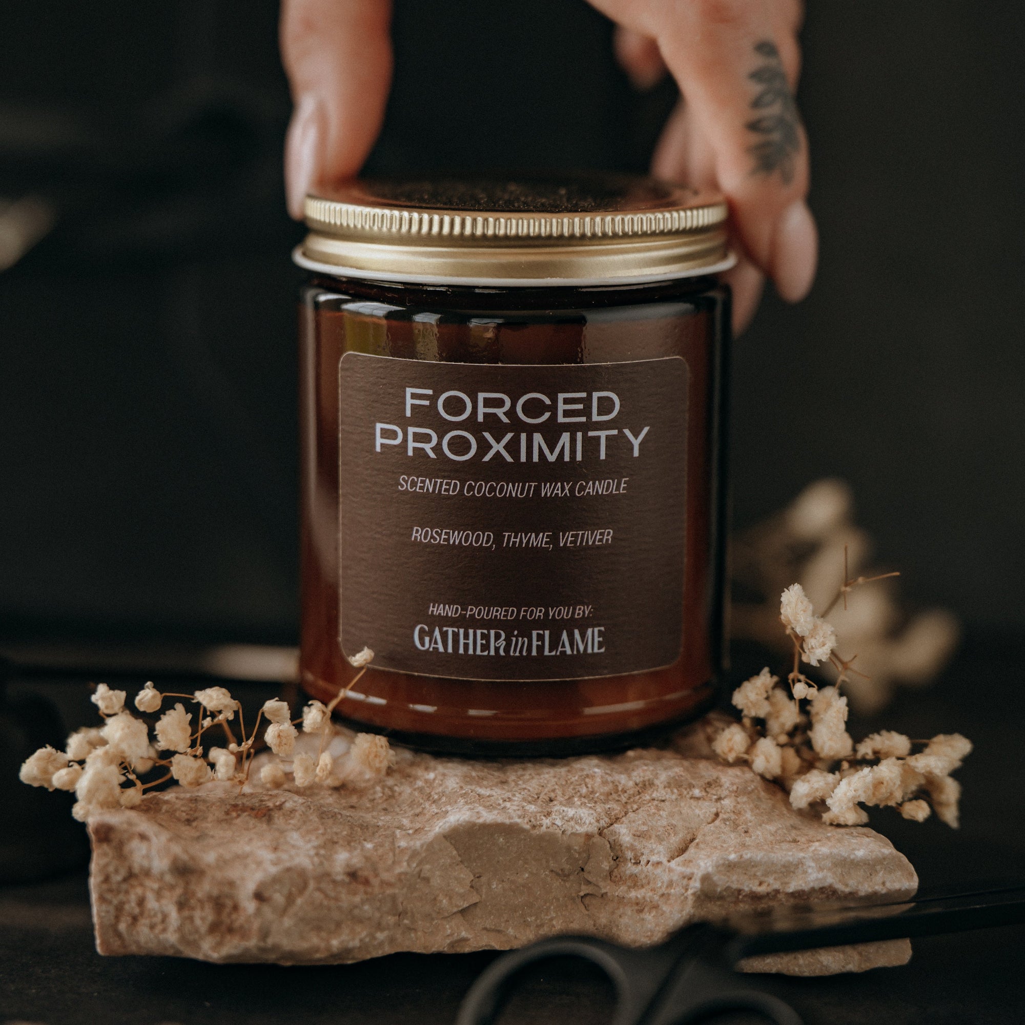 Jar of 'Forced Proximity' candle by Gather and Flame on a dark background with dried flowers and scissors.