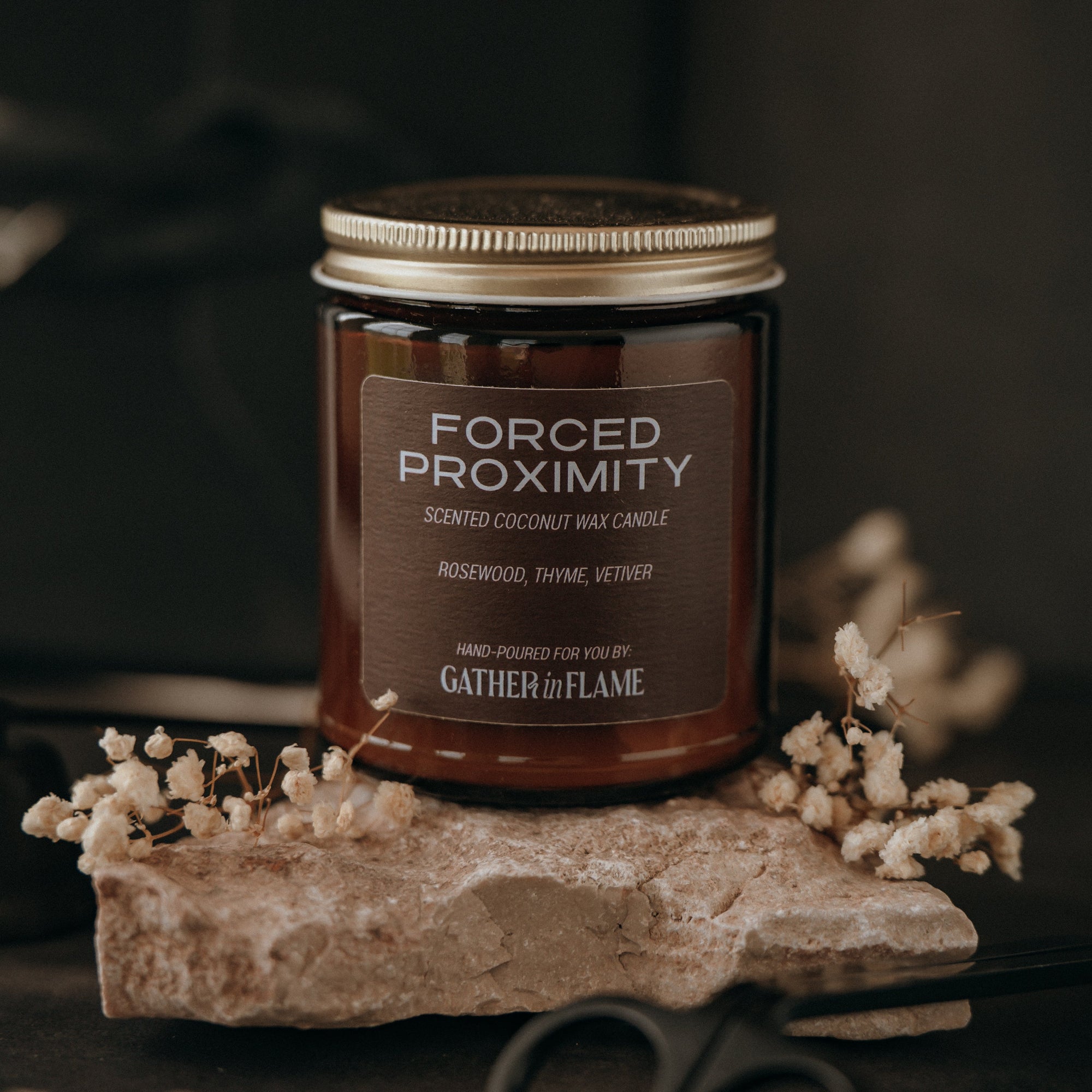 Candle jar labeled 'Forced Proximity' on a stone surface with flowers and scissors in the background.
