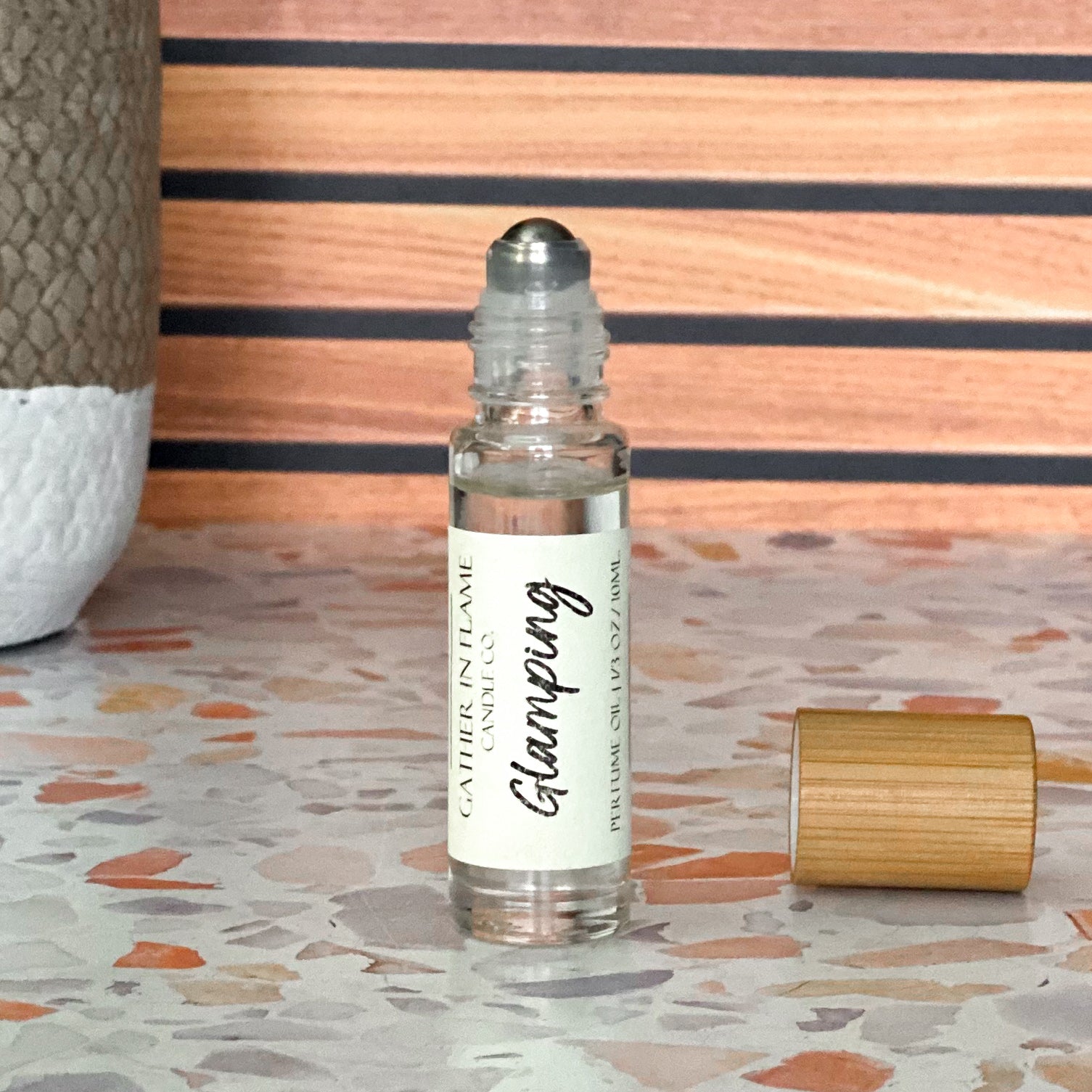 A perfume oil roller with a clear glass bottle and a bamboo cap sitting beside it, labeled 'Glamping', resting on a terrazzo surface with wood background.