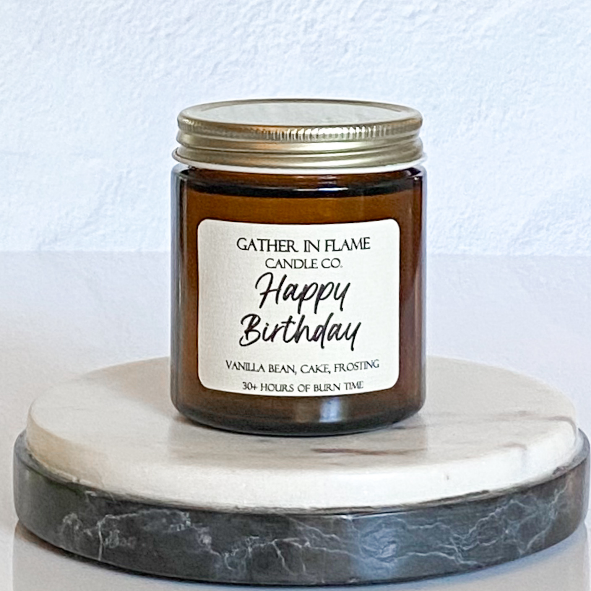 A jar candle with a gold lid, white label with 'Happy Birthday' text, and a description of the scent 'Vanilla Bean Cake Baking'. The candle is placed on a round, grey marble coaster.