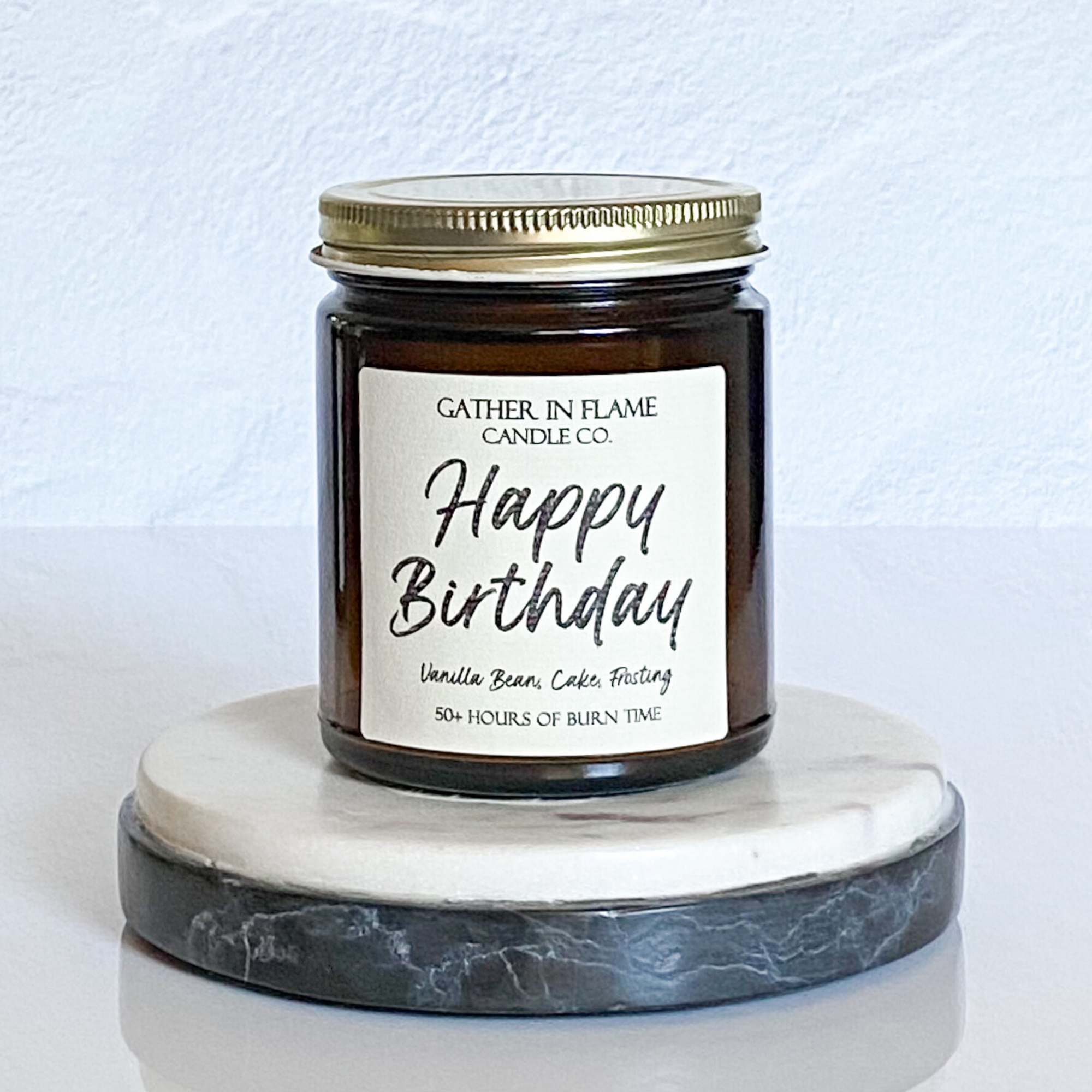 A jar candle with a gold lid, white label with 'Happy Birthday' text, and a description of the scent 'Vanilla Bean Cake Frosting'. The candle is placed on a round, grey marble coaster.