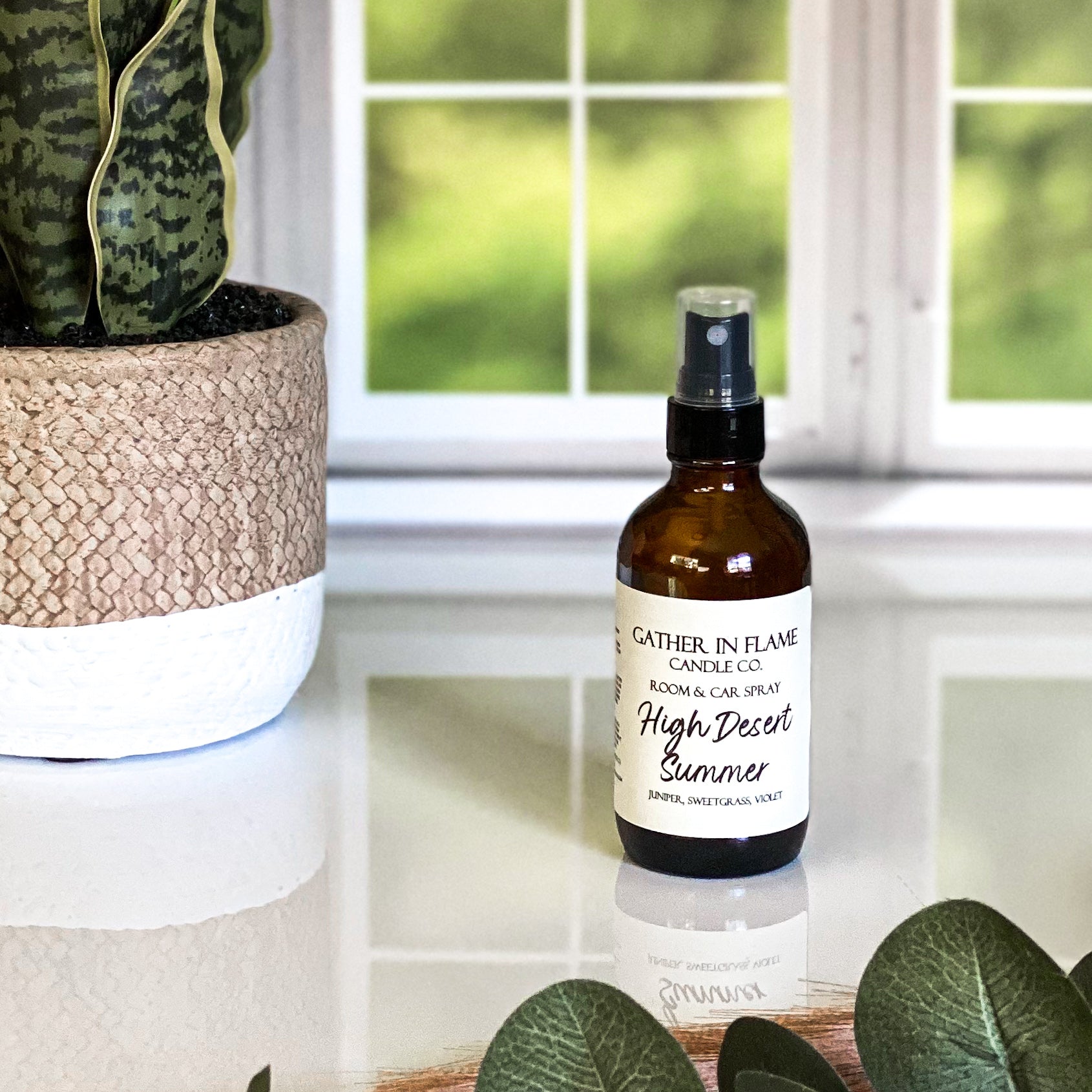 A brown amber spray bottle labeled 'High Desert Summer' by Gather In Flame Candle Co. on a white marble surface with a window and plant in the background.