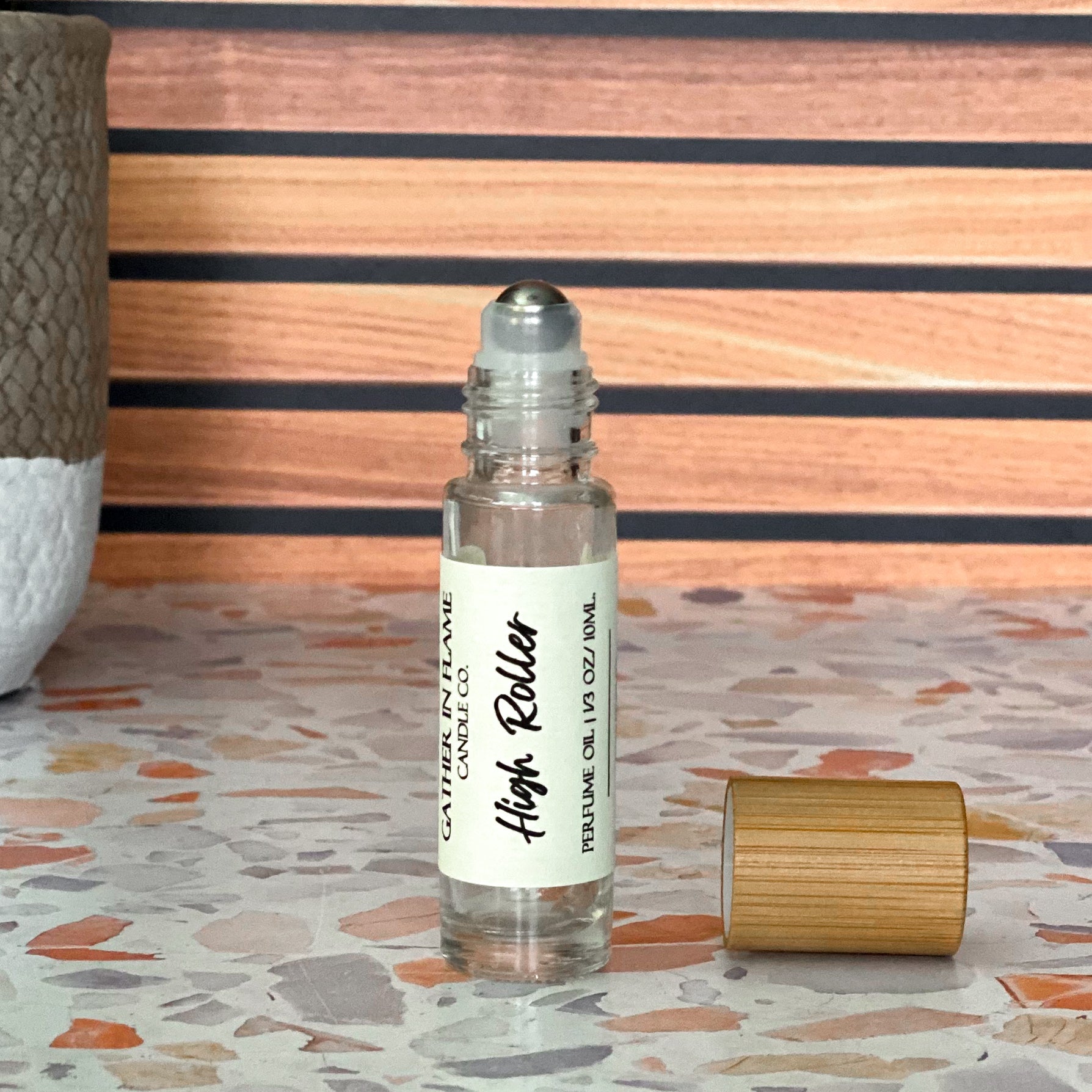 A clear perfume oil roller with a bamboo cap sitting beside it, labeled 'High Roller' on a terrazzo surface with wood background.