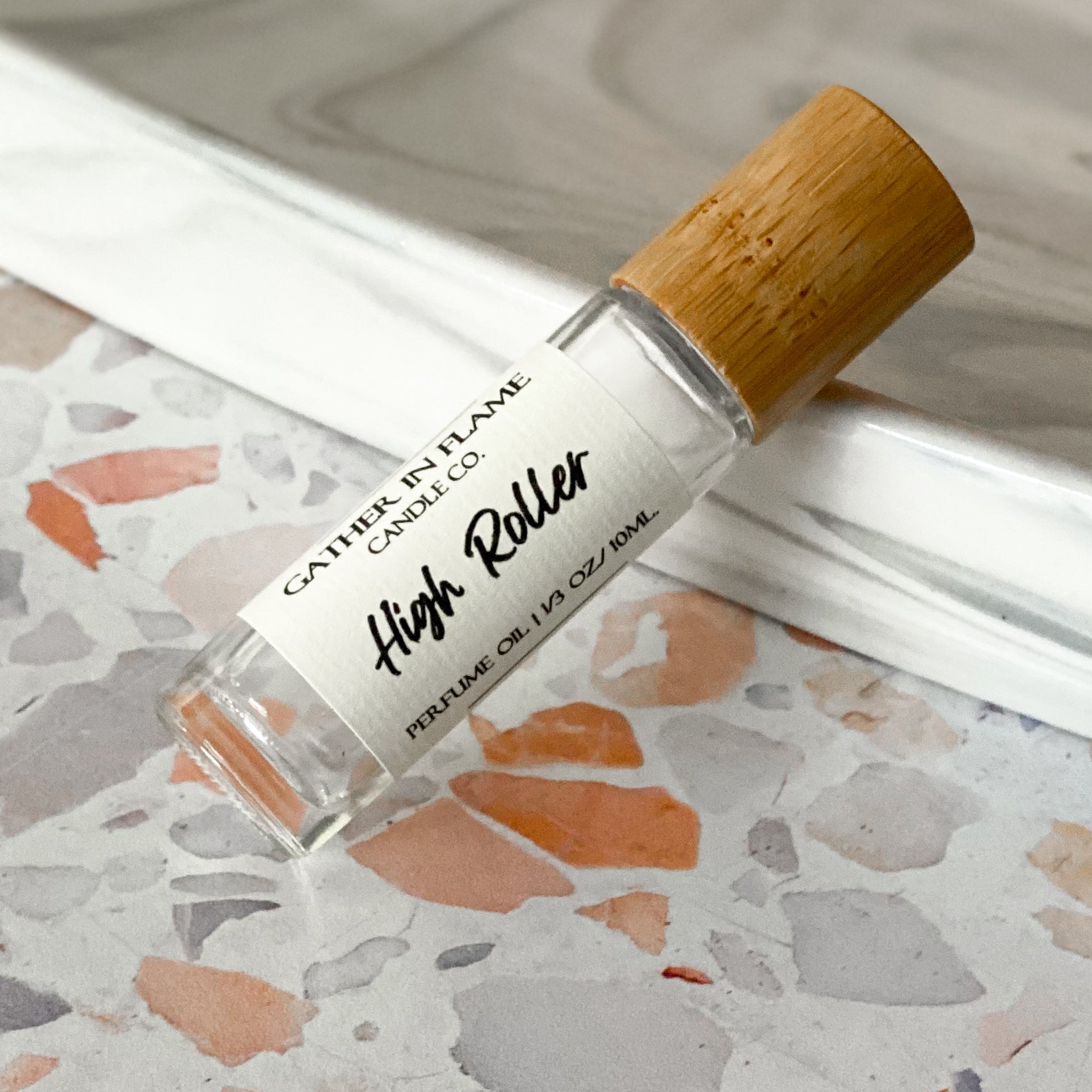 A clear perfume oil roller with a bamboo cap labeled 'High Roller' on a terrazzo surface.