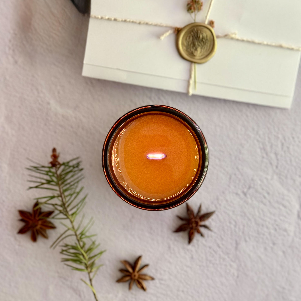 Topdown view of lit candle with wood wick surrounded by star anise, pine needles and a handwritten letter with wax seal on a textured surface