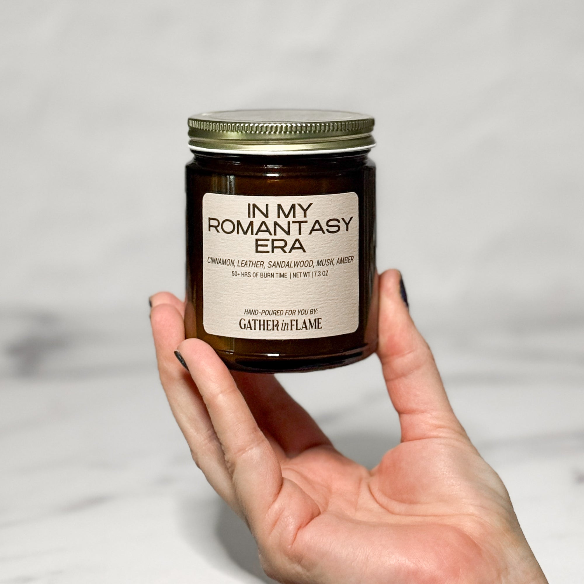 Amber glass jar candle labeled In My Romantasy Era held in a hand with a white background.