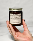 Amber glass jar candle labeled In My Romantasy Era held in a hand with a white background.