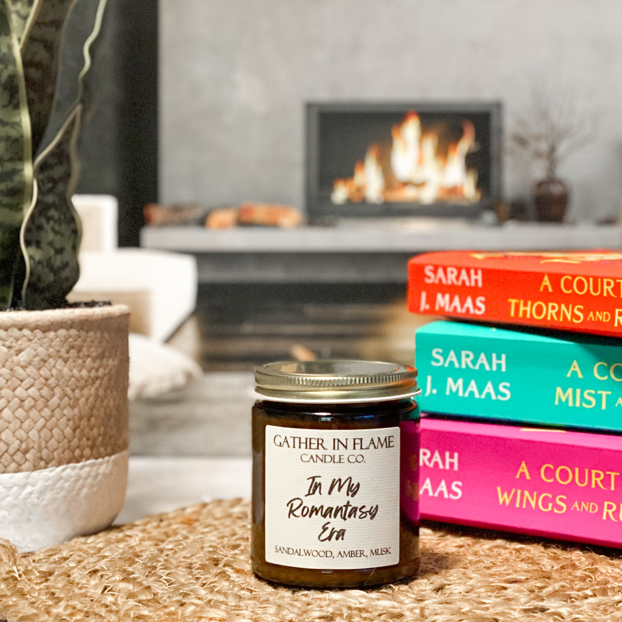 A jar candle with a white label that reads 'In My Romantasy Era', placed on a woven surface with books and a plant in the background.