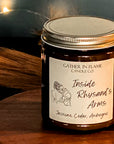 Close up of a  jar candle with a gold lid, labeled 'Inside Rhysand's Arms', depicting an illustration of a female character and a male character, with woodsy and floral scent notes. On a wood surface with fairy lights, pampas grass and blue in the background.