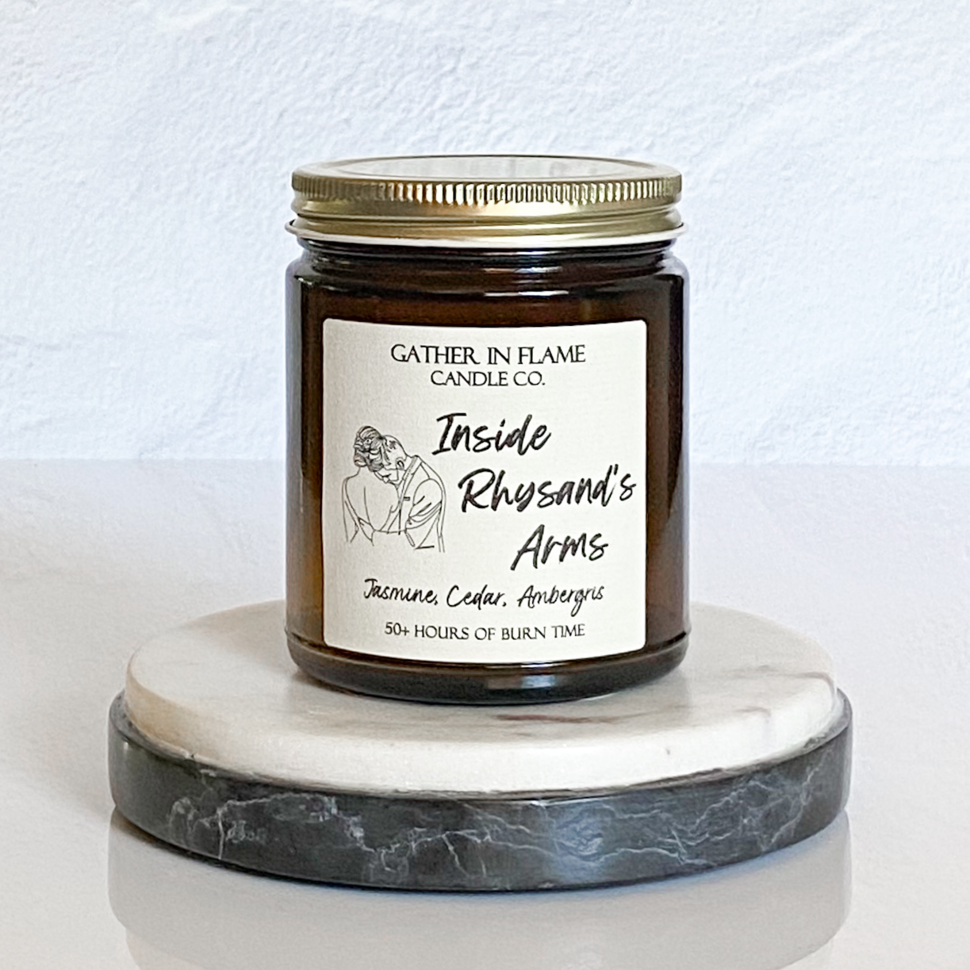 A jar candle with a gold lid, labeled 'Inside Rhysand's Arms', depicting an illustration of a female character and a male character, with woodsy and floral scent notes.