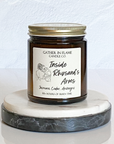 A jar candle with a gold lid, labeled 'Inside Rhysand's Arms', depicting an illustration of a female character and a male character, with woodsy and floral scent notes.