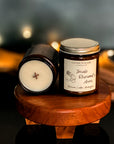 Jar candle with a gold lid, labeled 'Inside Rhysand's Arms', depicting an illustration of a female character and a male character, with woodsy and floral scent notes next to a candle surface showing smooth top and wooden x wick. On a wood surface with fairy lights, pampas grass and blue in the background.
