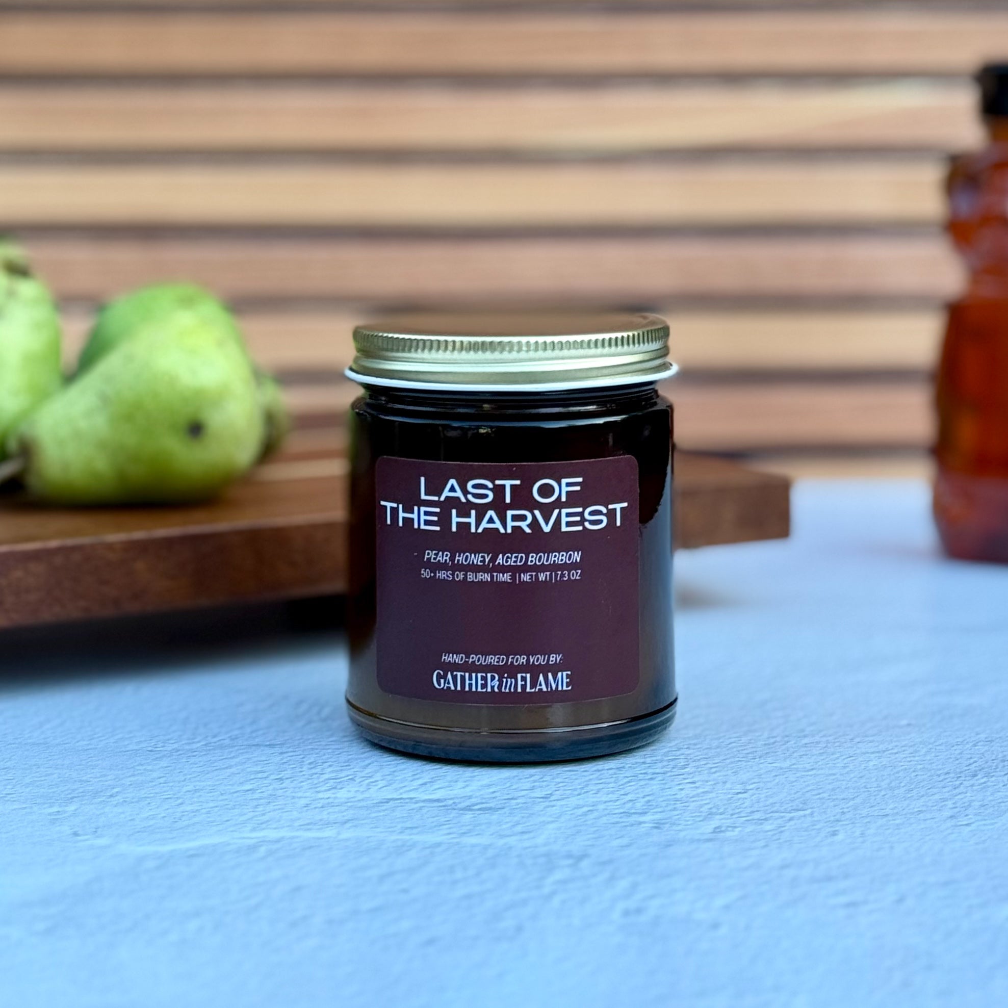 Candle jar labeled &#39;Last of the Harvest&#39; by Gather the Flame on a blue surface with pears and a bottle of honey in the background.