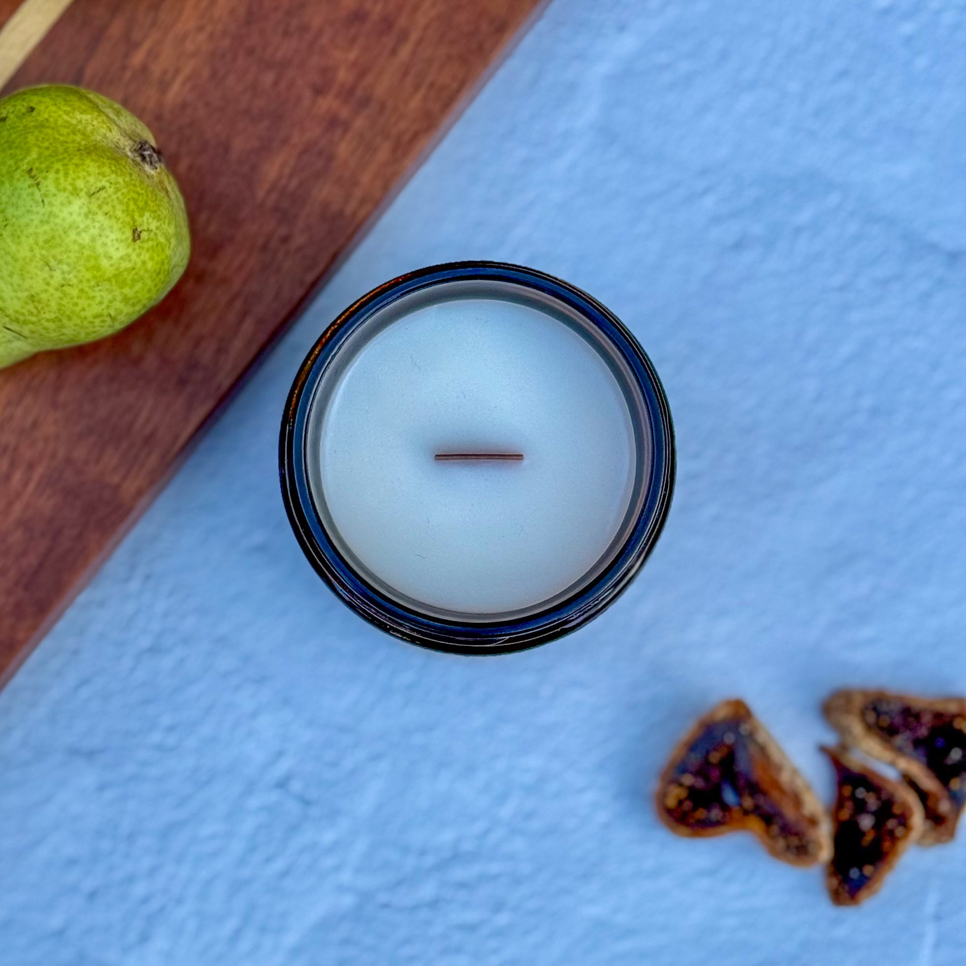 Topdown view of candle with wood wick on a textured blue surface with a pear and fig slices.