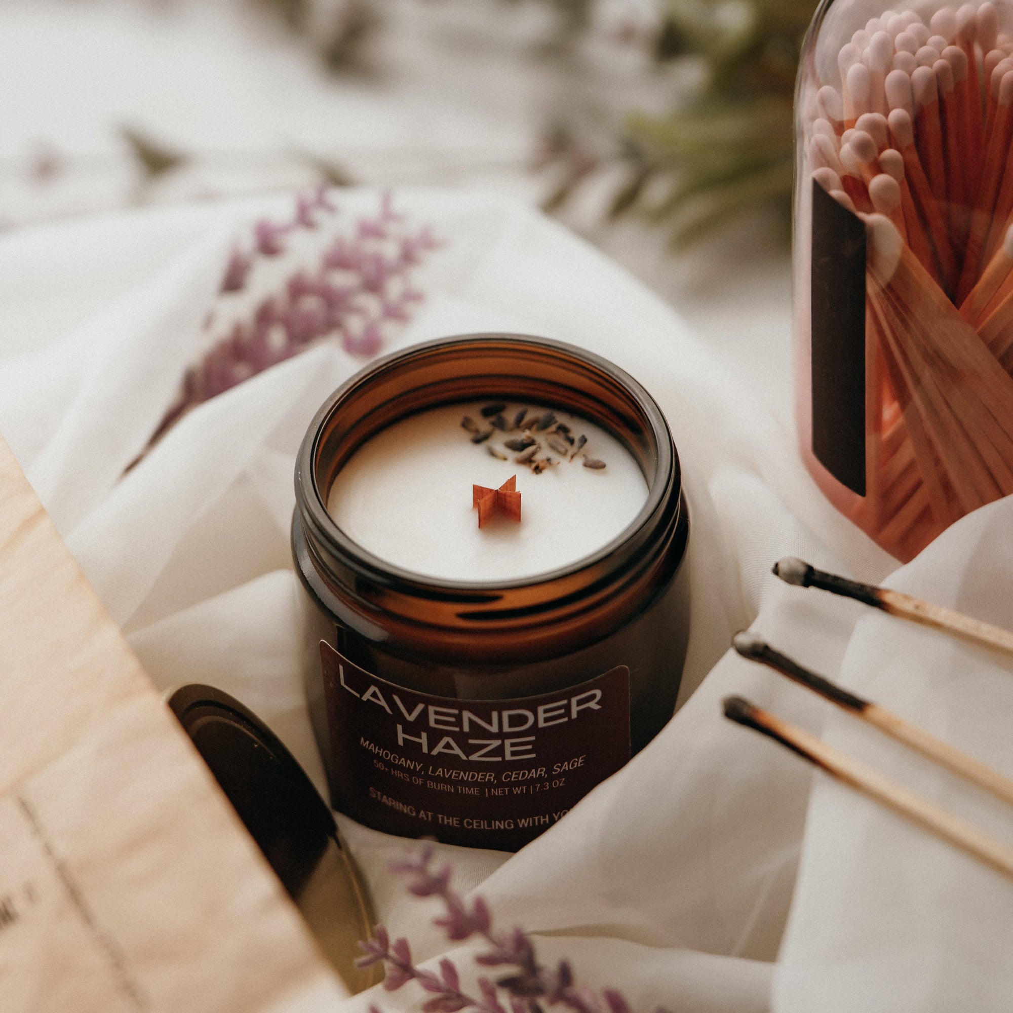 Lavender-scented candle in a jar with matches and a book on a soft surface
