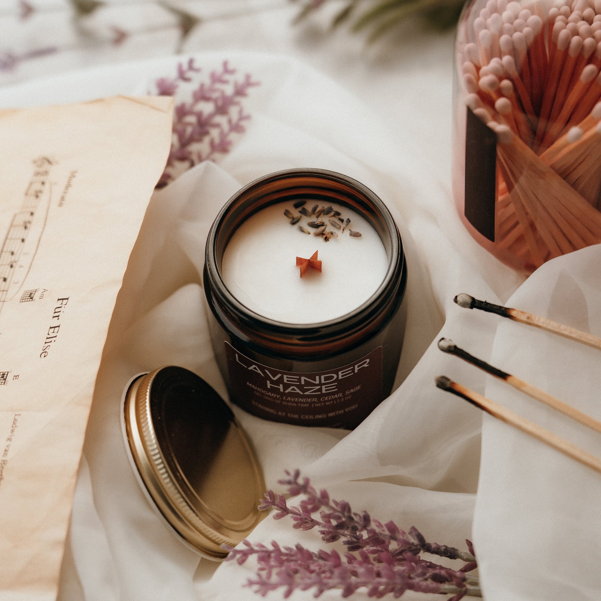 Candle in a jar with lavender scent on a white surface with matches and a book.