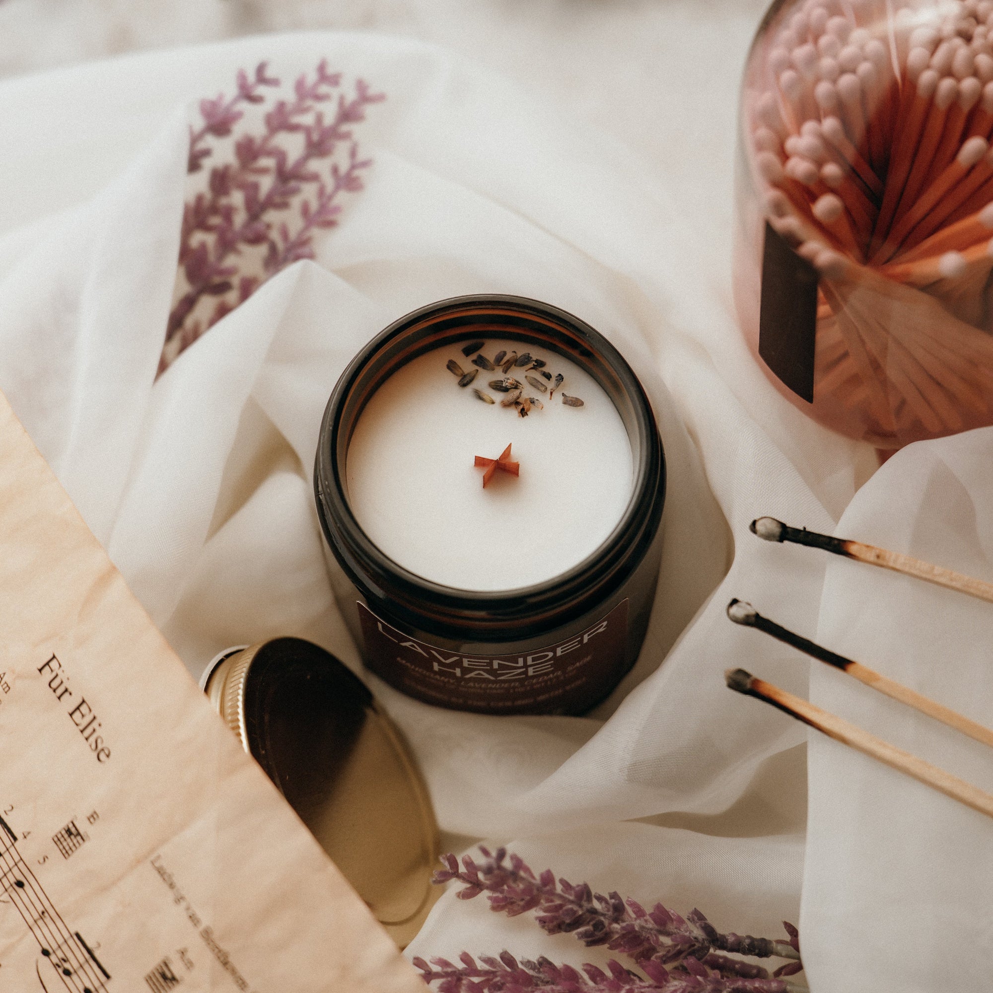 Candle in a black container with lavender flowers, surrounded by a book and matchsticks on a white surface.