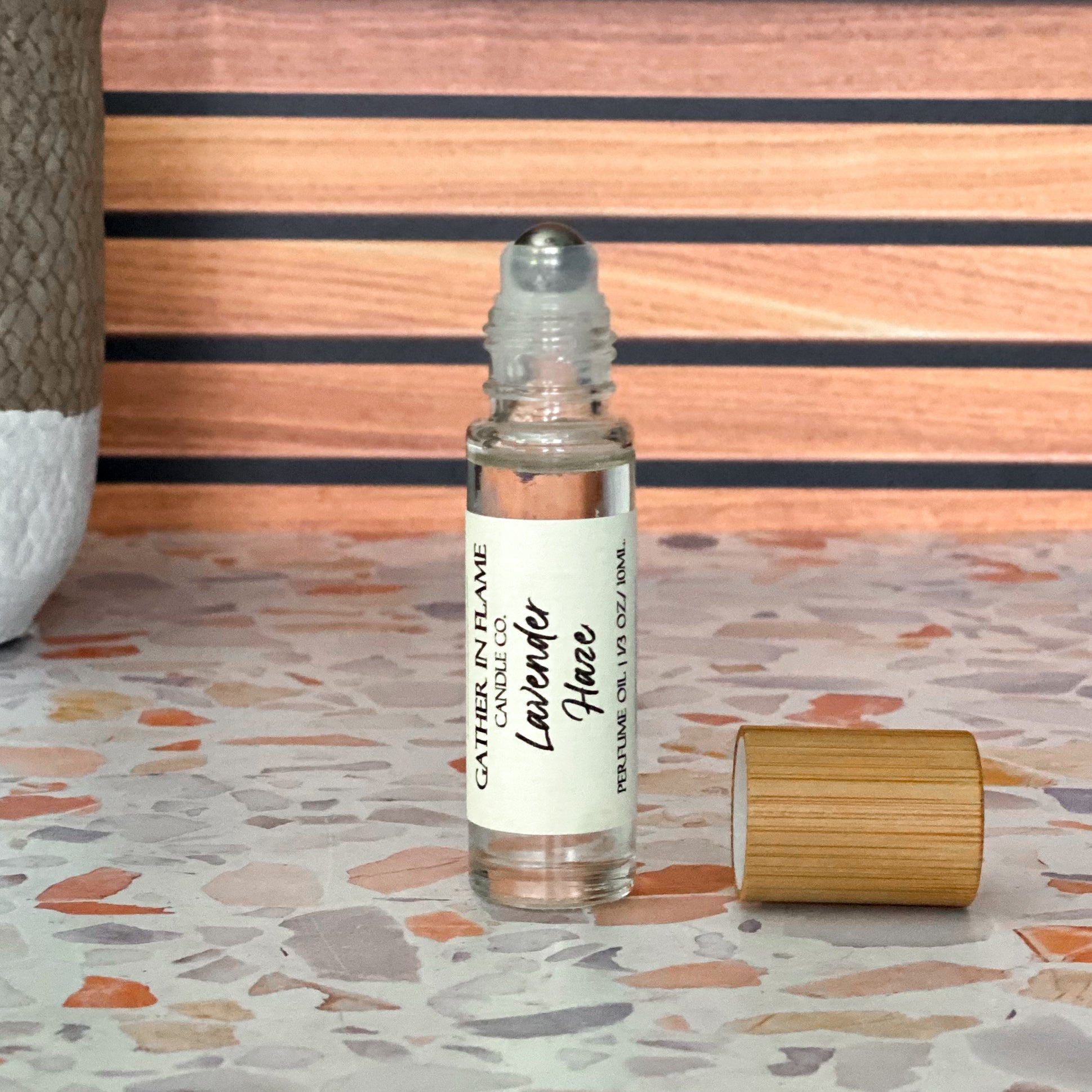 A perfume oil roller with a clear glass body and a bamboo cap sitting beside it, labeled 'Lavender Haze' on a terrazzo surface with wood background.