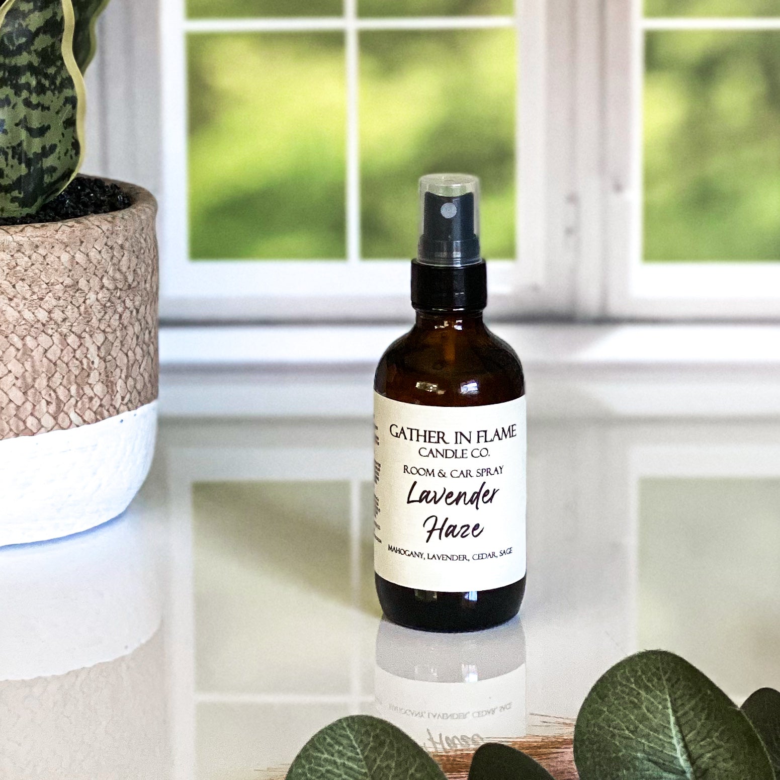 A brown amber spray bottle labeled 'Lavender Haze' by Gather In Flame Candle Co. on a white marble surface with a window and plant in the background.