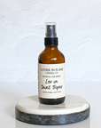 A brown amber spray bottle labeled 'Gather In Flame Candle Co. - Room & Car Spray - Leo in Saint Tropez' placed on a white marble surface.