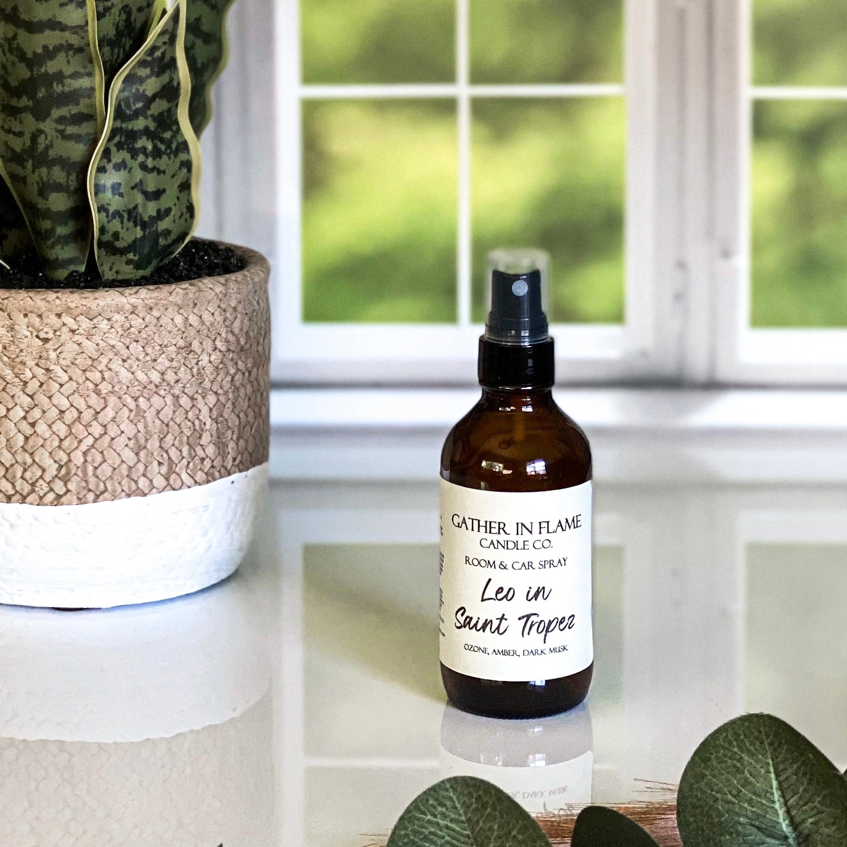 A brown amber spray bottle labeled 'Leo in Saint Tropez' by Gather In Flame Candle Co. on a white marble surface with a window and plant in the background.