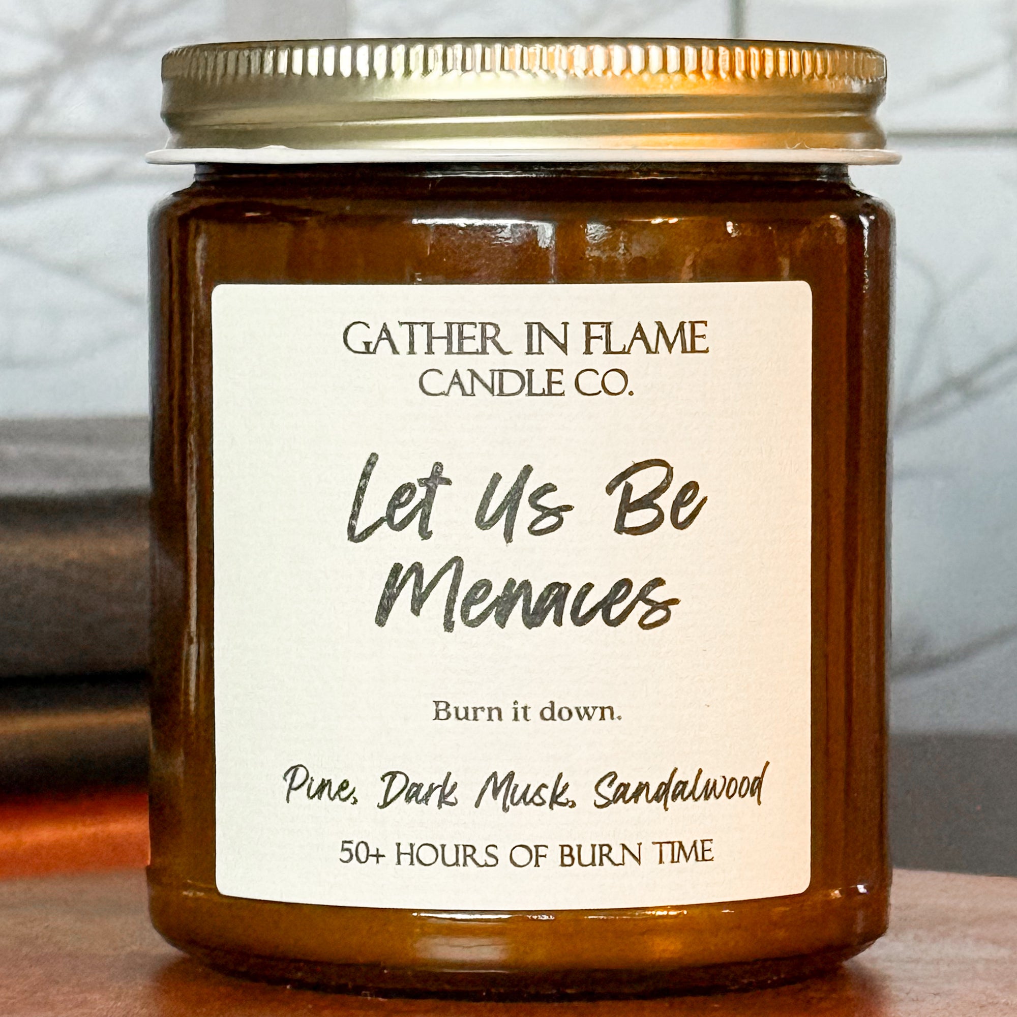 Close up of a brown jar candle with a gold lid, labeled 'Let Us Be Menaces', placed on a wooden stand, with books and a window in the background.