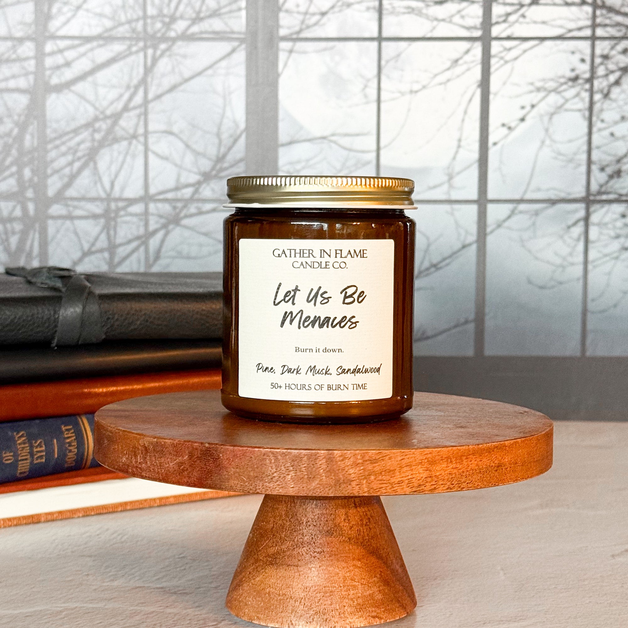 A brown jar candle with a gold lid, labeled 'Let Us Be Menaces', placed on a wooden stand, with books and a window in the background.