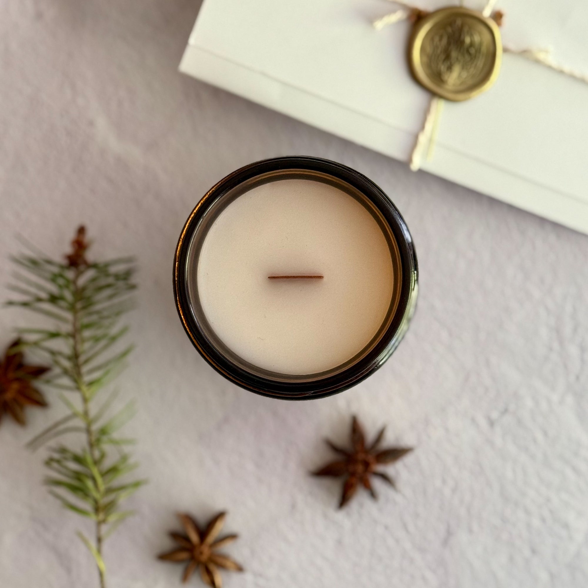Topdown view of candle with wood wick surrounded by star anise, pine needles and a handwritten letter with wax seal on a textured surface