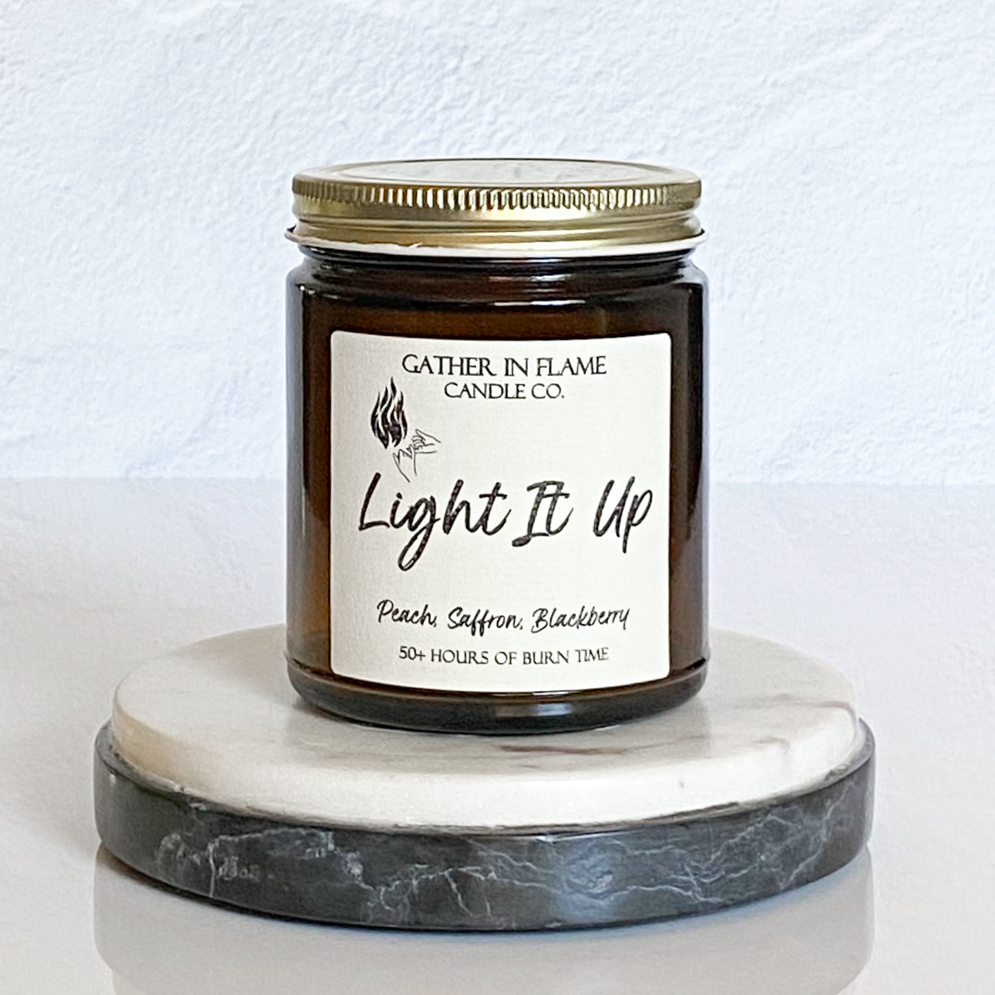 A jar candle with a gold lid, labeled 'Light It Up' by Gather In Flame Candle Co., with scents of peach, saffron, blackberry, and amber.