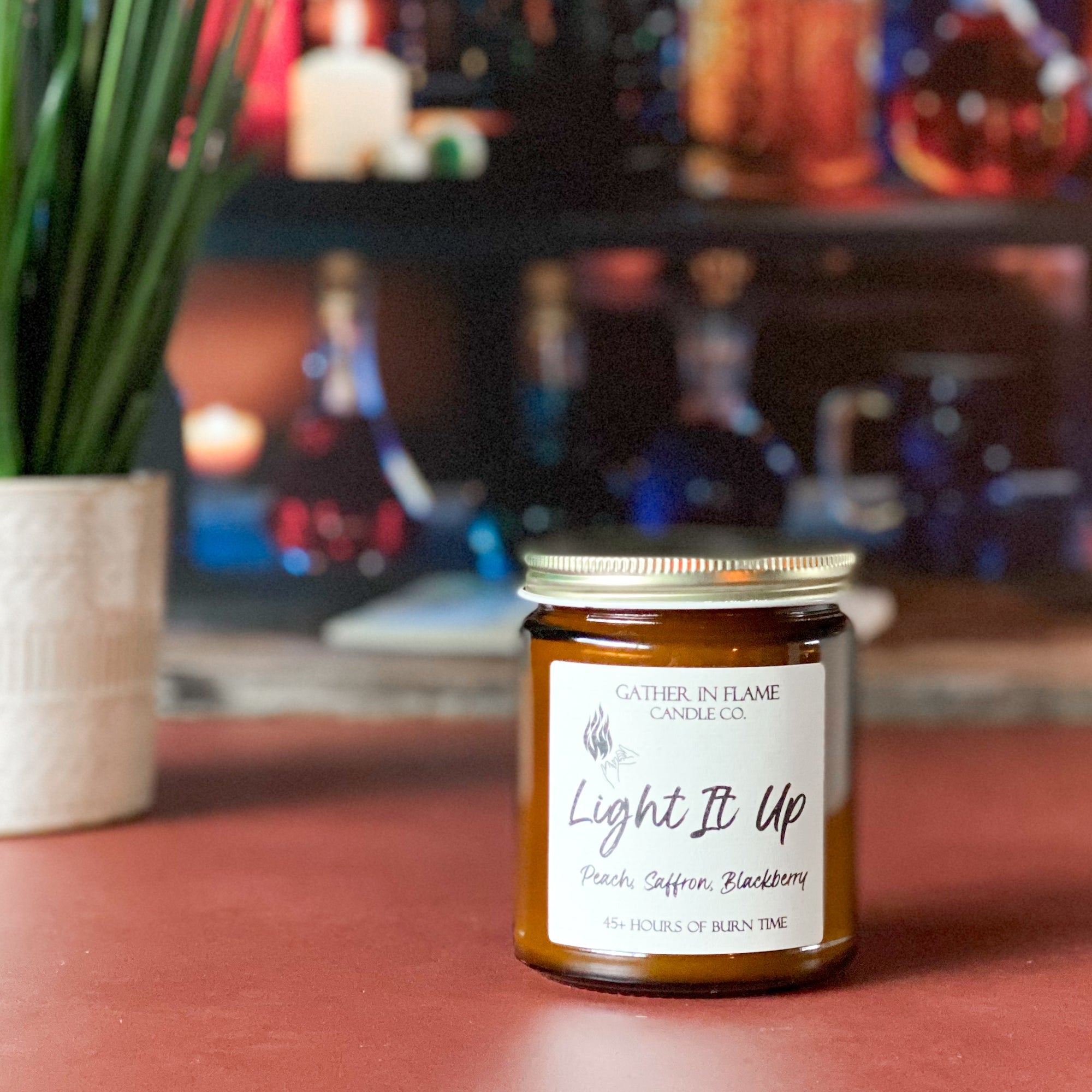 A jar candle with a gold lid, labeled 'Light It Up' by Gather In Flame Candle Co., with scents of peach, saffron, blackberry, and amber on an orange surface with bottles in the background.