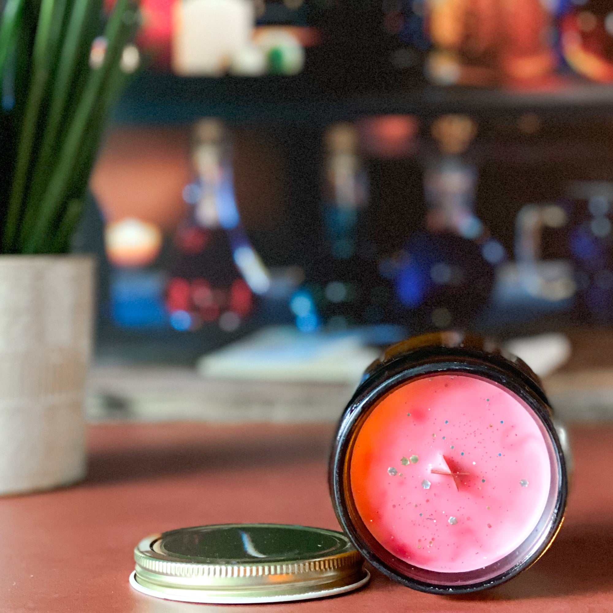 The surface of a candle with reddish color and gold glitter with a gold lid on an orange surface with a plant and bottles in the background.