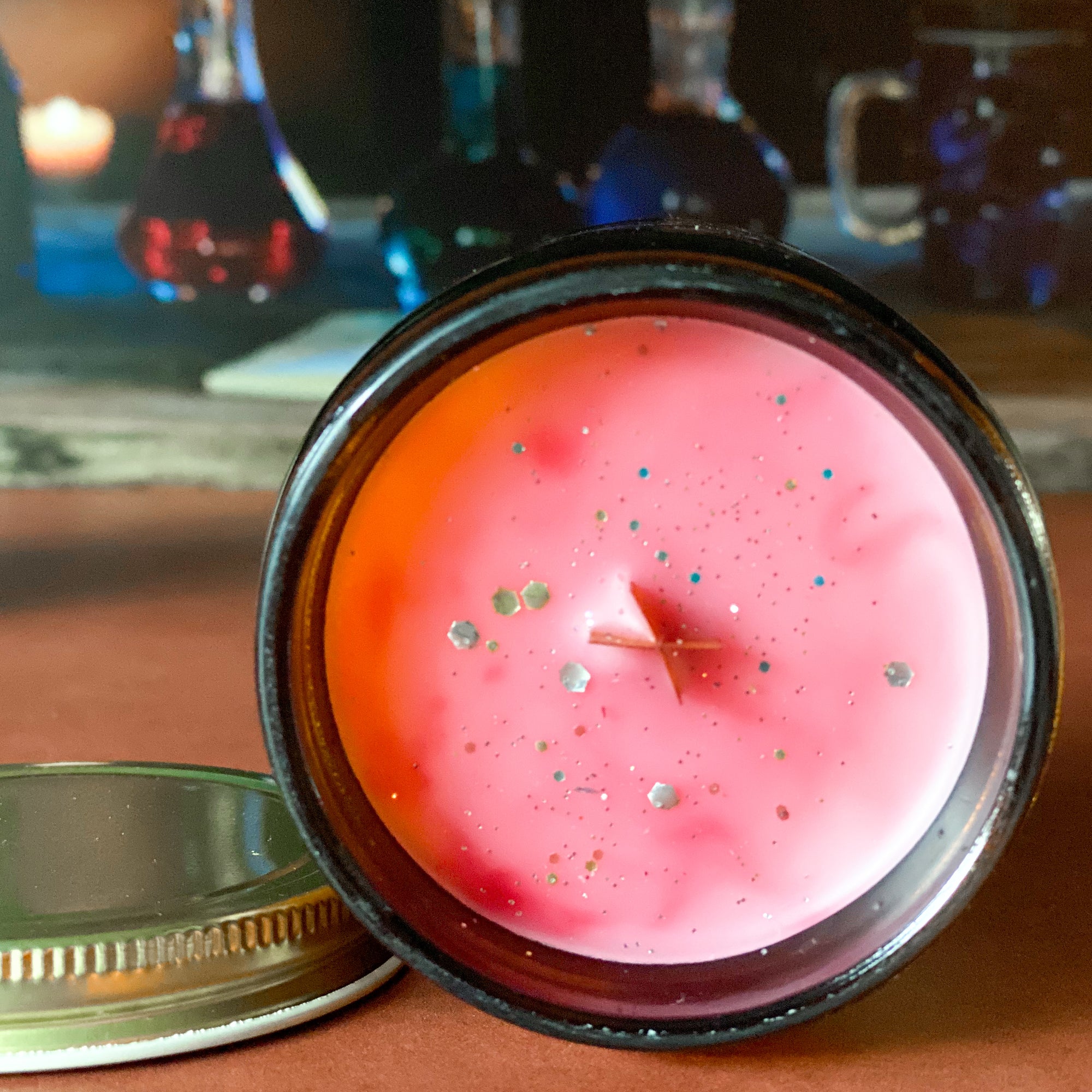The surface of a candle with reddish color and gold glitter on an orange surface with bottles in the background.
