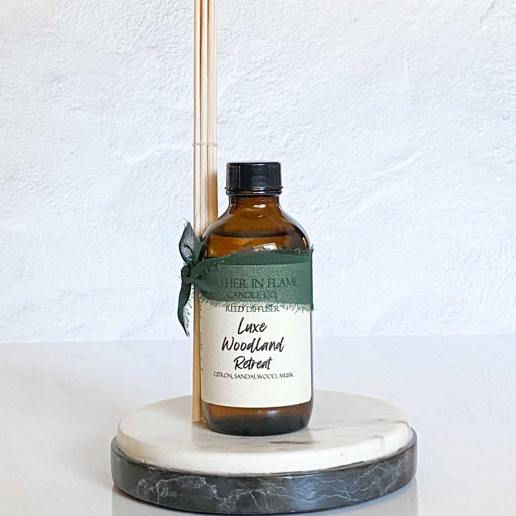 A brown reed diffuser bottle with a label reading 'Luxe Woodland Retreat', set on a white marble round tray with a green ribbon decoration.