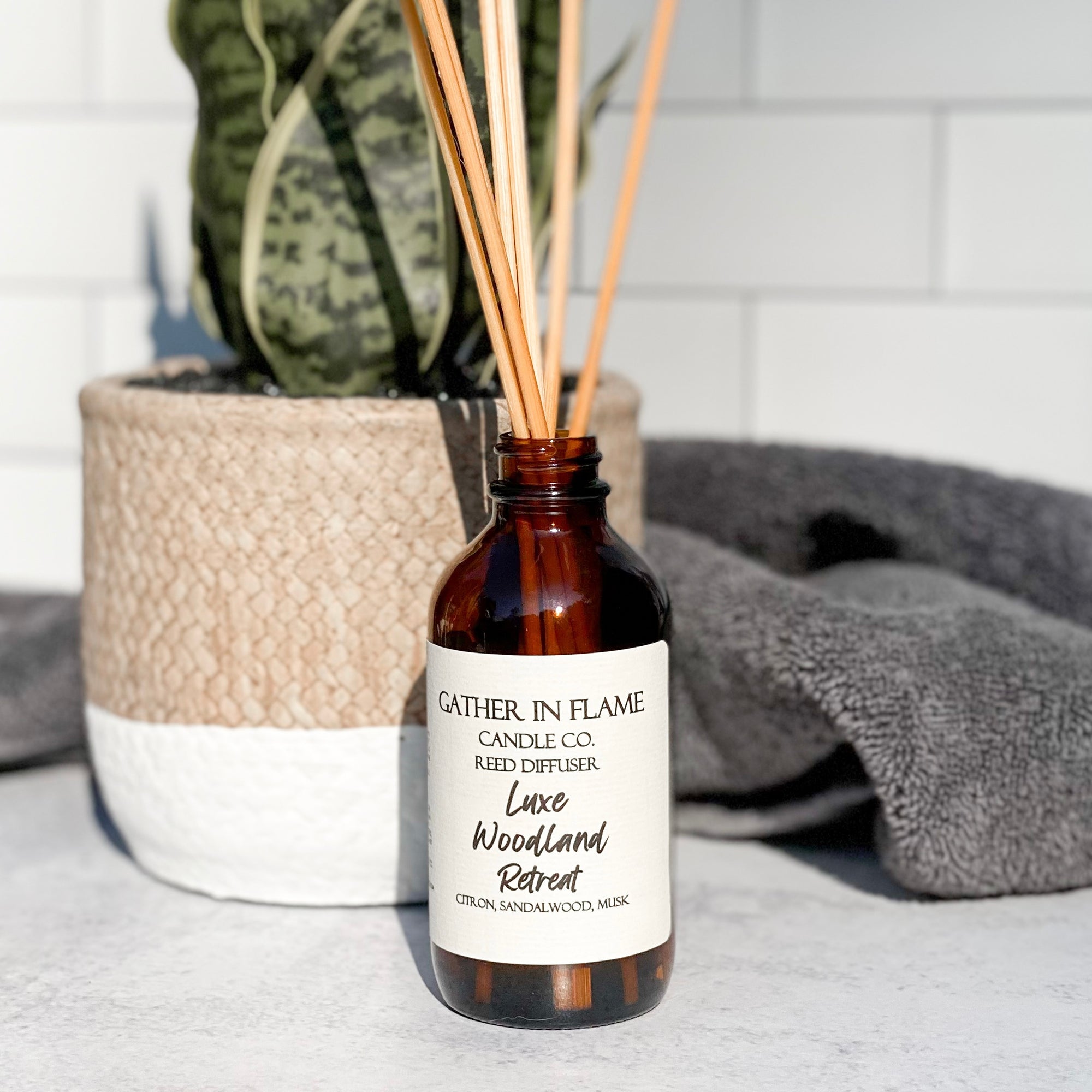 A brown glass reed diffuser bottle with a label reading "Luxe Woodland Retreat", placed on a grey surface, surrounded by plant and towels with white tile background.