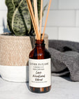 A brown glass reed diffuser bottle with a label reading "Luxe Woodland Retreat", placed on a grey surface, surrounded by plant and towels with white tile background.